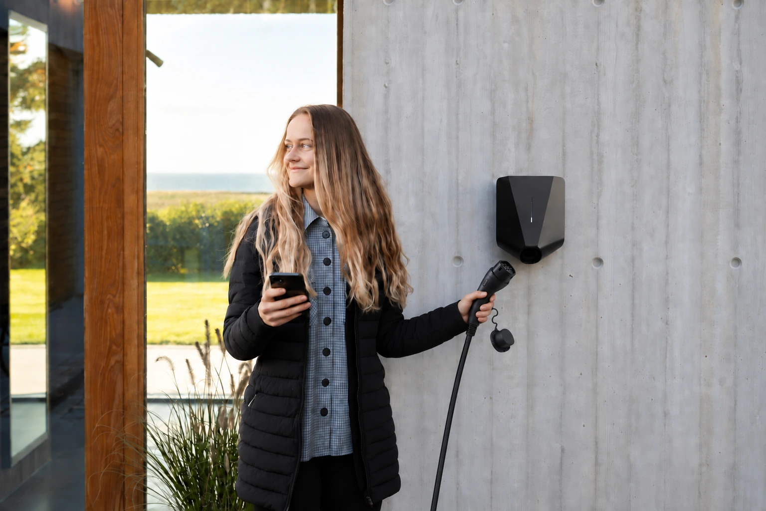 Illustration : une femme tient son téléphone et le câble devant une borne Easee noire sur un mur en béton